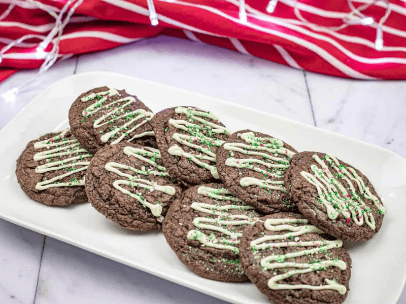 A plate of chocolate cookies topped with white icing and green sprinkles. A red and white striped cloth is in the background.