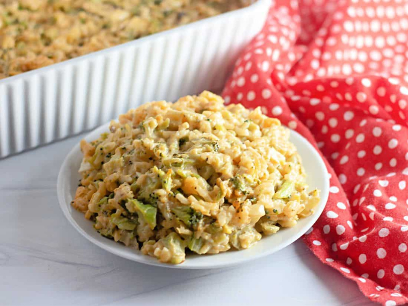 A serving of broccoli rice casserole on a white plate, with the casserole dish and a red polka dot cloth in the background.