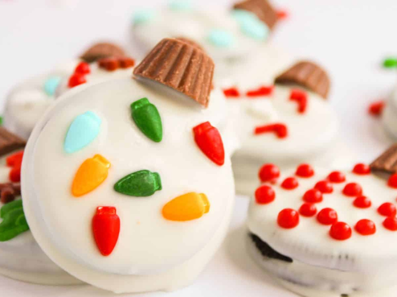 A close-up of Oreo Christmas Ornament Cookies, elegantly coated in white chocolate and adorned with colorful candy lights, red sprinkles, and chocolate pieces on top.