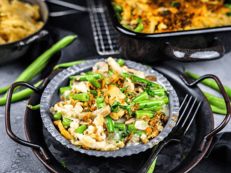 A plate of green bean casserole with mushrooms and fried onions sits on a tray, with a fork and green beans nearby.