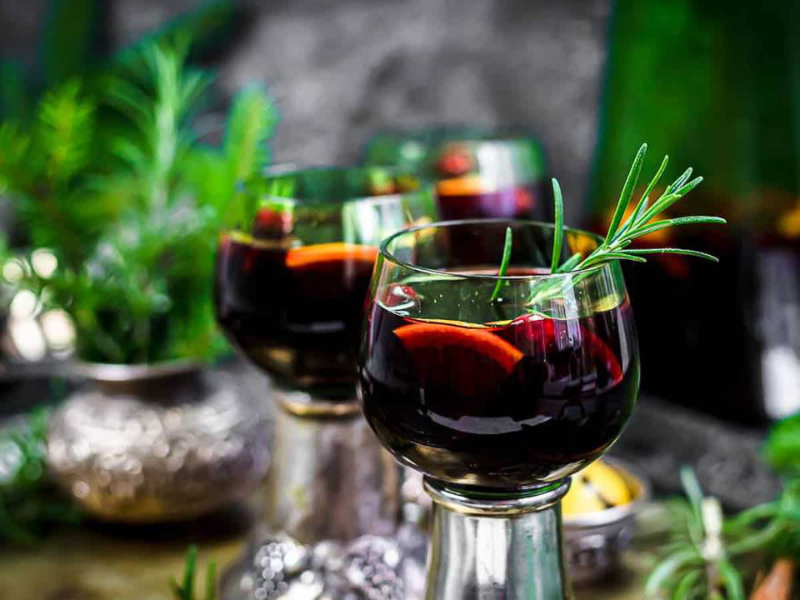 Three glasses of red wine with orange slices and rosemary sprigs, set on a table with greenery and a blurred background.