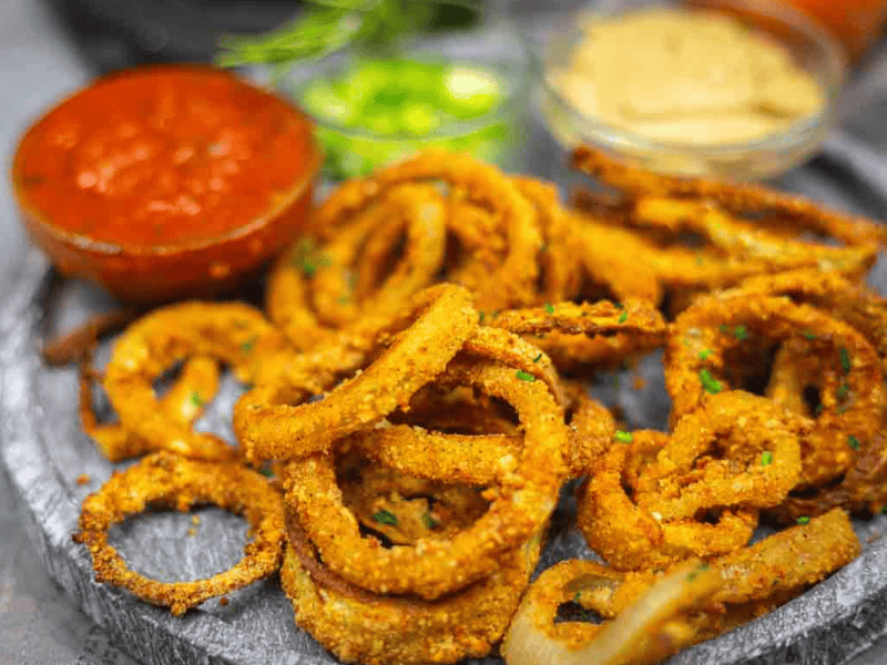 A plate of crispy fried onion rings with three dipping sauces, including marinara and mustard, served on a round gray tray.