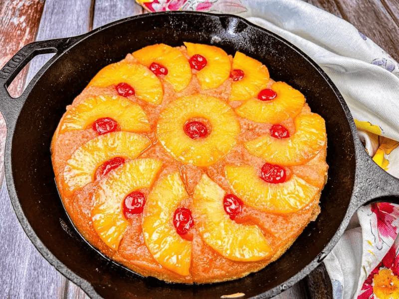 A pineapple upside-down cake with pineapple rings and maraschino cherries is baked in a black cast iron skillet on a wooden surface.
