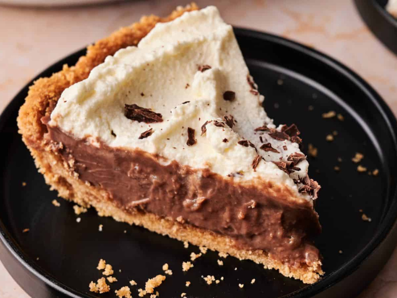 A slice of chocolate cream pie with a graham cracker crust, topped with whipped cream and chocolate shavings, served on a black plate.