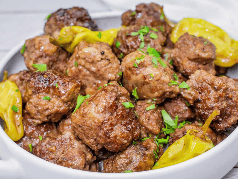 A bowl of browned meatballs garnished with chopped parsley and served with yellow peppers.