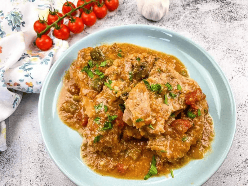 A plate of meat stew with vegetables and sauce, garnished with chopped herbs, sits on a light blue plate. Cherry tomatoes and a garlic bulb are in the background.