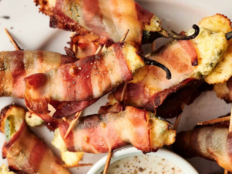 Bacon-wrapped stuffed jalapeño peppers arranged on a plate with toothpicks, next to a small bowl of dipping sauce.