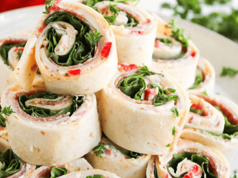 A plate of tortilla pinwheel sandwiches filled with spinach, cream cheese, red peppers, and herbs, neatly stacked and garnished with parsley.