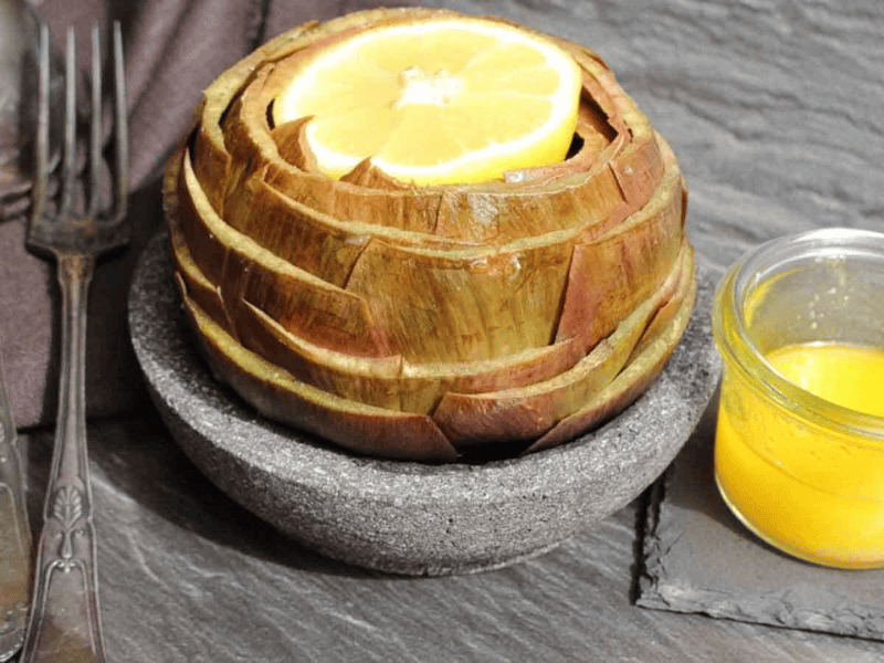 A cooked artichoke with a lemon slice on top sits in a bowl, next to a fork and a small jar of yellow dipping sauce on a stone surface.