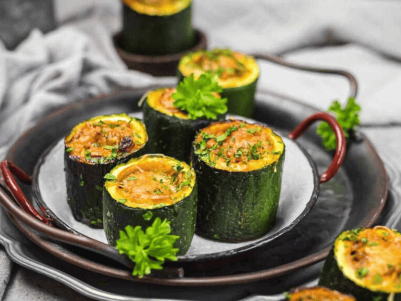 Six baked zucchini cups filled with a golden, cheesy mixture are served on a dark plate, garnished with parsley.