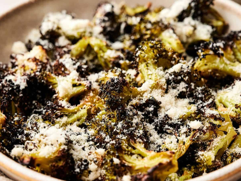 A close-up of roasted broccoli florets topped with grated Parmesan cheese in a ceramic bowl.