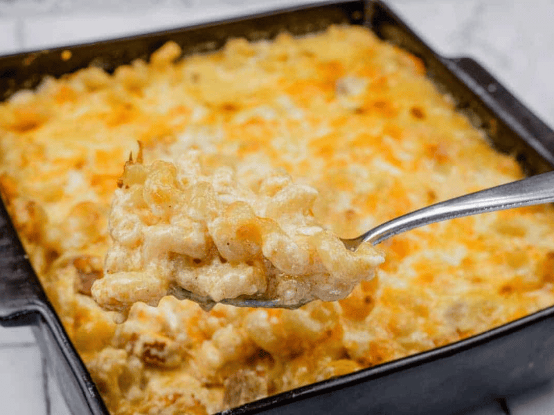 A spoonful of Tini’s Mac & Cheese with a Twist is held above a square baking dish filled with the same dish.