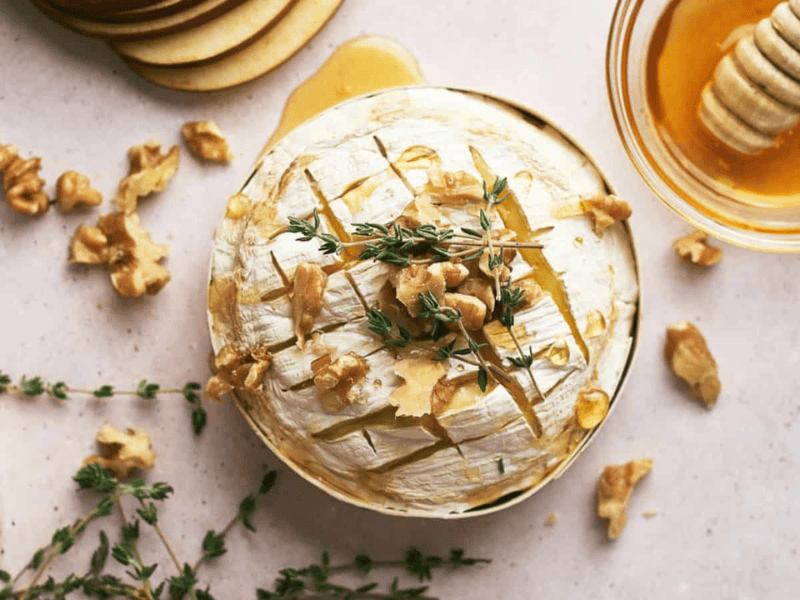 Round baked brie cheese topped with walnuts, fresh thyme, and drizzled honey, served in a wooden container.