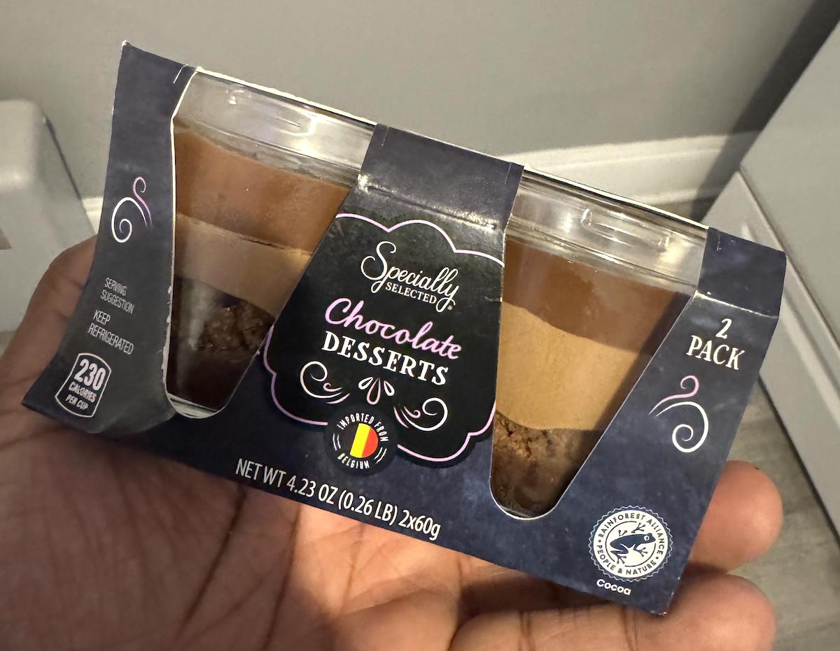 A hand holds a two-pack of Specially Selected Chocolate Desserts, each in a clear cup, showing chocolate layers. The label notes 230 calories per serving and UTZ Certified cocoa.