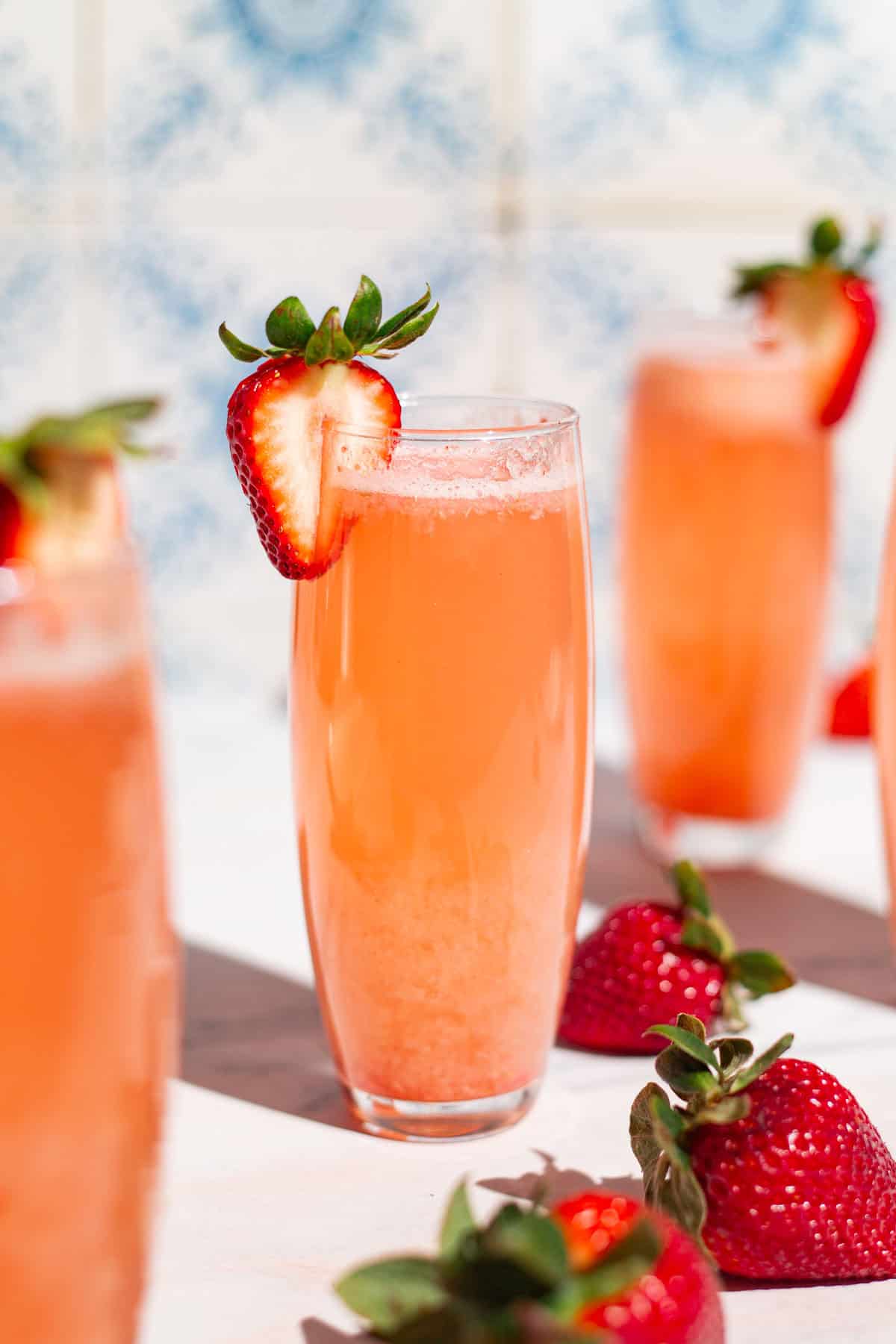A close up of a strawberry bellini in a glass garnished with a strawberry slice surrounded by more glasses of the bellini and whole strawberries.