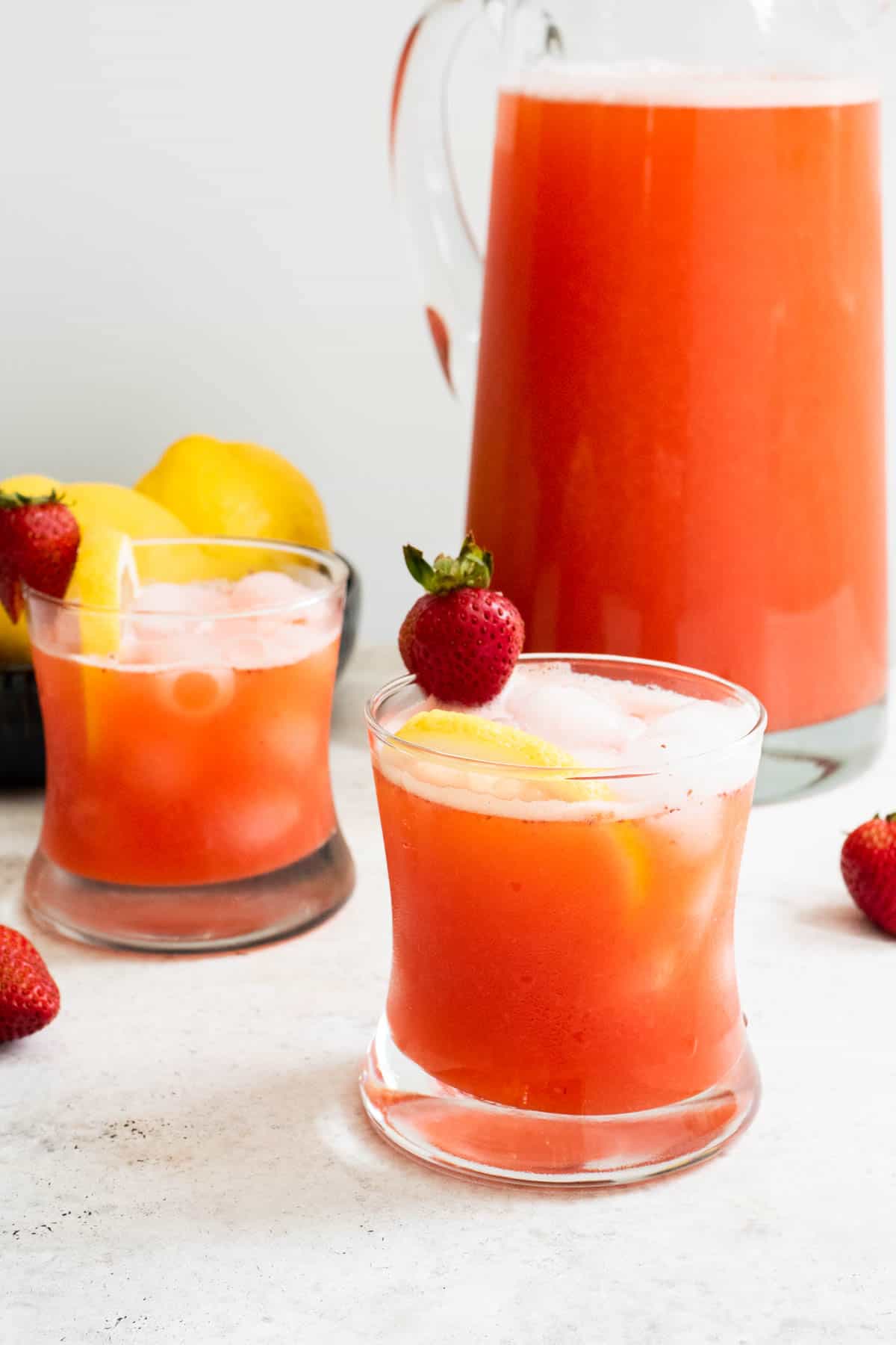 two glasses of strawberry lemonade garnished with strawberries and lemon slices in front of a pitcher of strawberry lemonade.