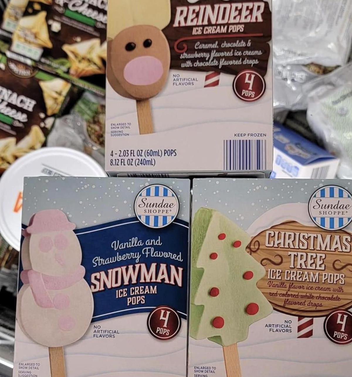 Three boxes of Sundae Shoppe holiday ice cream pops—Reindeer, Snowman, and Christmas Tree varieties—are displayed in a grocery store freezer. Each box features four pops in festive shapes and flavors.
