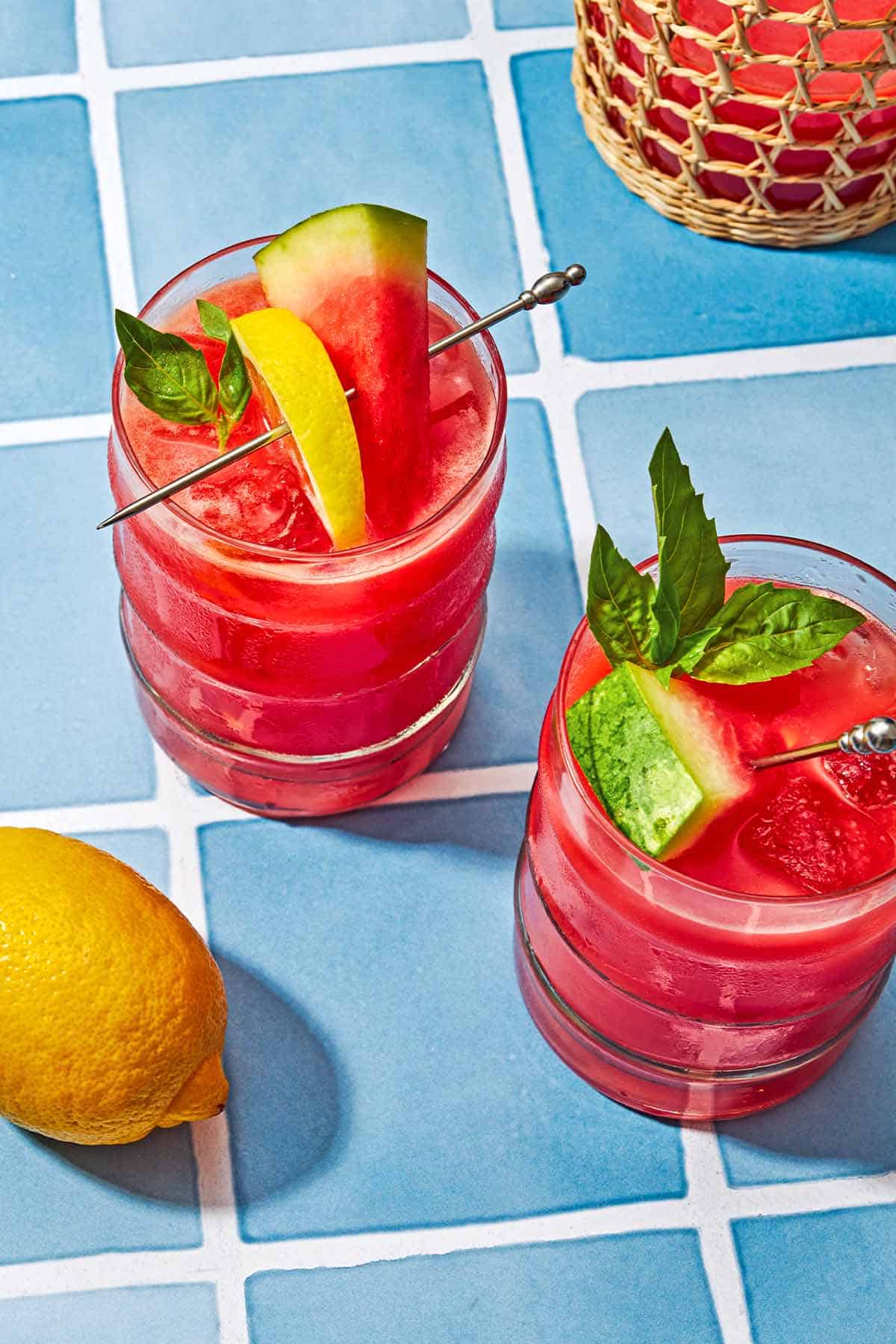 Two glasses of watermelon lemonade garnished with basil and slices of watermelon and lemon next to a whole lemon.