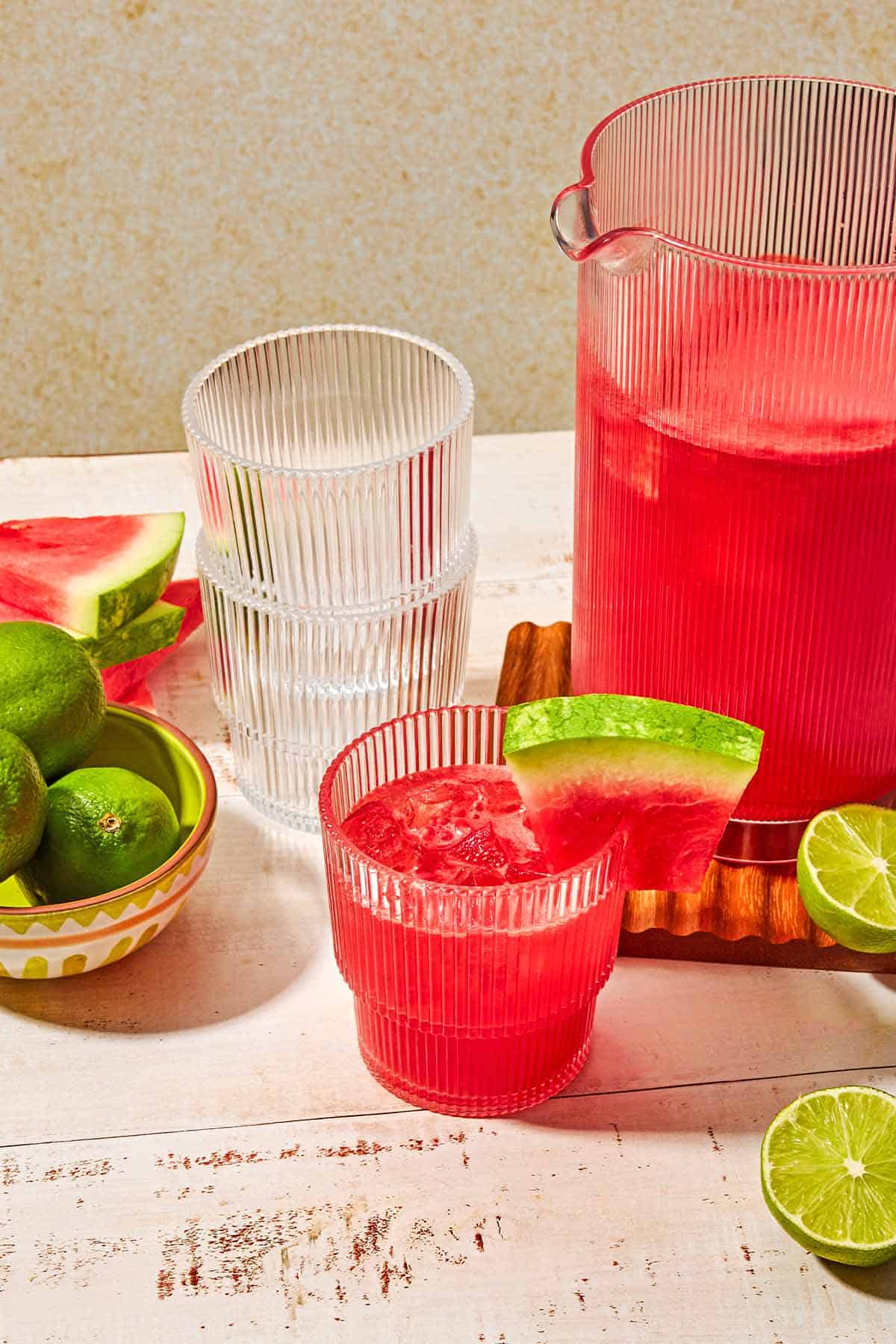 A pitcher of watermelon sangria next to 2 lime halves, a bowl of limes, a stack of watermelon slices, and empty glass and a glass of the sangria garnished with a watermelon slice.