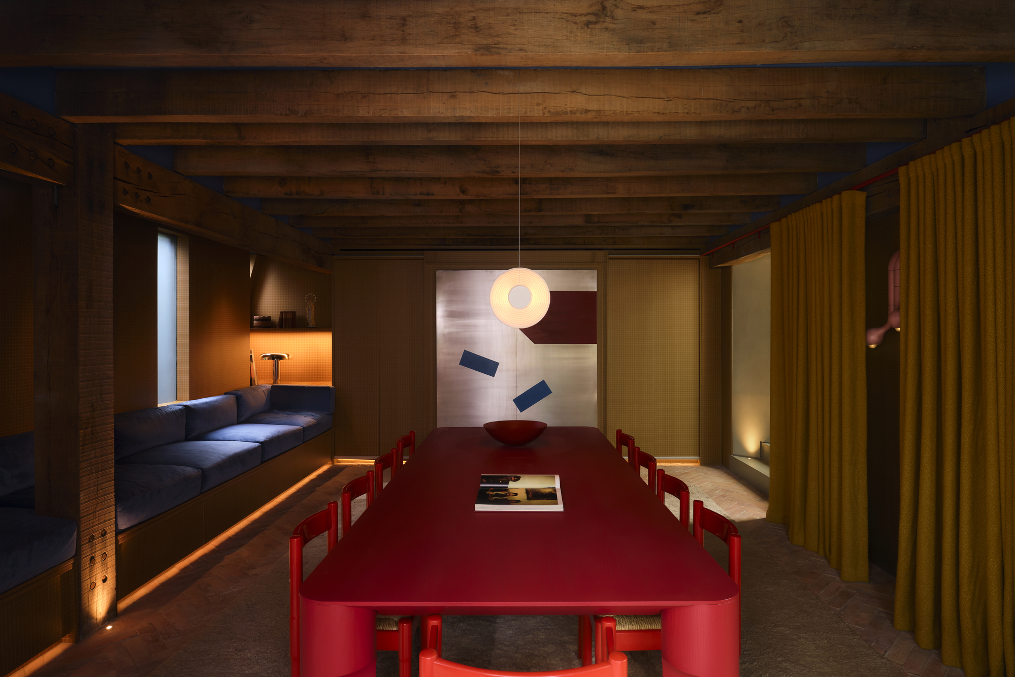 Image of a dark yellow dining room with dim lighting and wooden beams across the ceiling. There is a red dining table in the middle of the room and a navy blue bench seat to the side.
