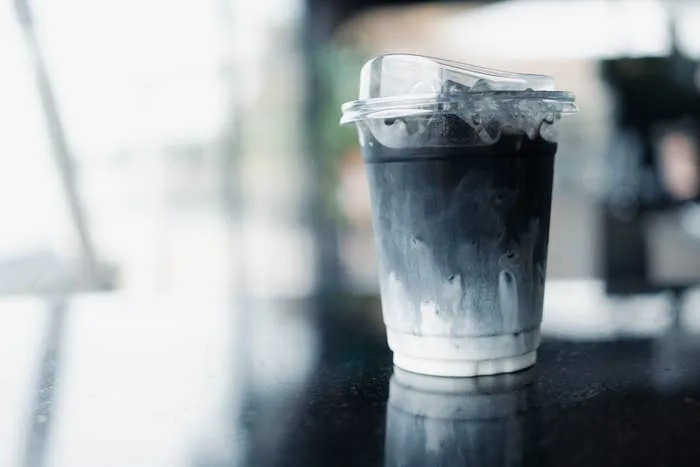 Icy black drink with swirls of white, served in a clear plastic cup with a lid, sitting on a reflective surface