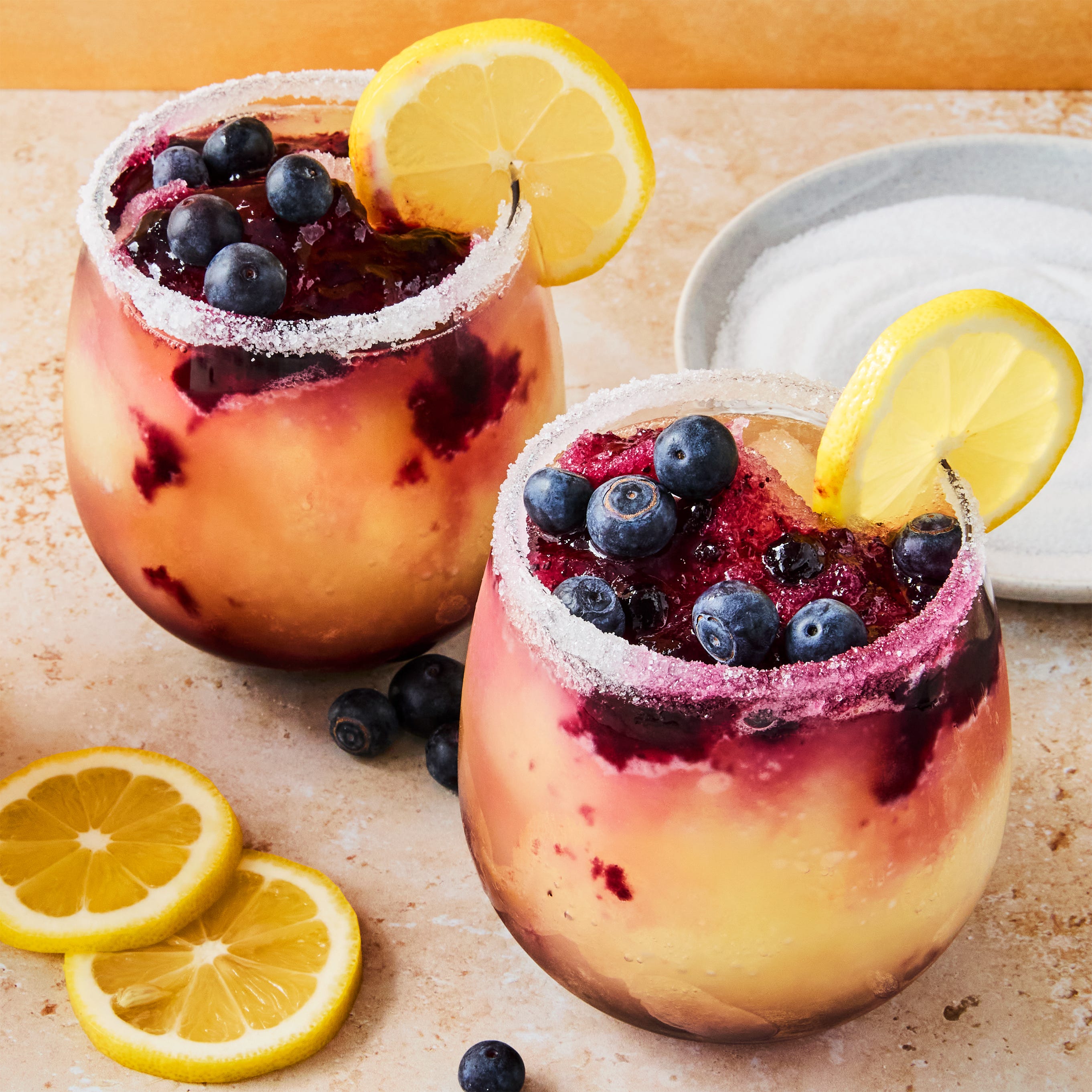stemless glasses filled with a frozen vodka lemon cocktail with a blueberry syrup swirled in the glass. garnished with a sugared rim, lemon wheels, and fresh blueberries