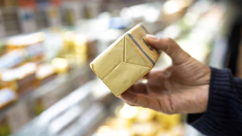 Close-up of a hand holding a wrapped block of butter in golden packaging at a grocery store