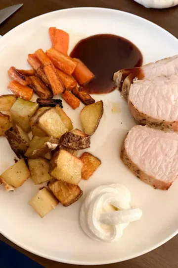 Sliced pork tenderloin with roasted carrots, potatoes, and a swirl of cream on a white plate.