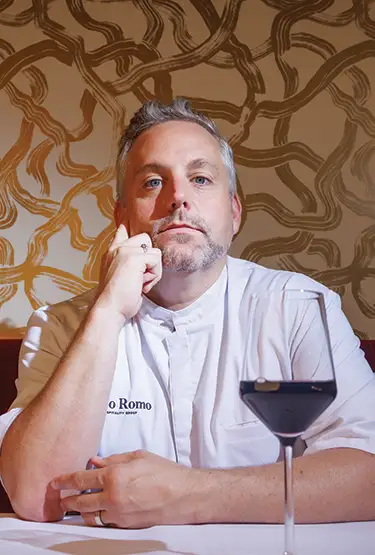 Chef Nico Romo poses at a table with a glass of wine.