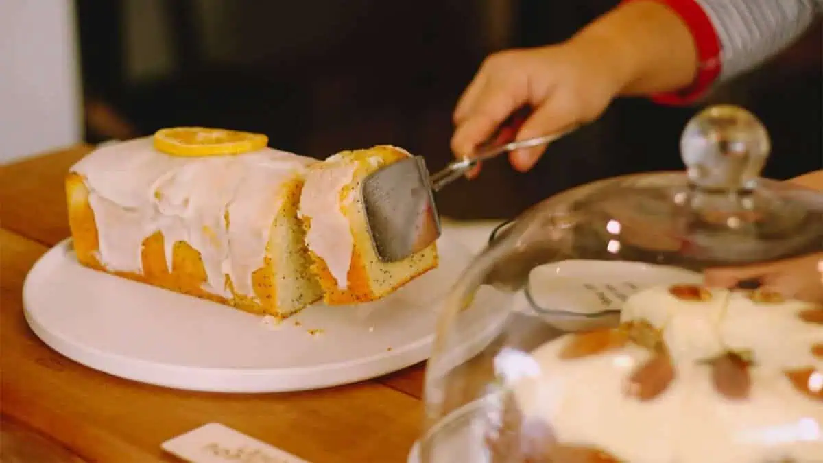 A hand slices a glazed loaf cake topped with a dried orange slice on a white platter.