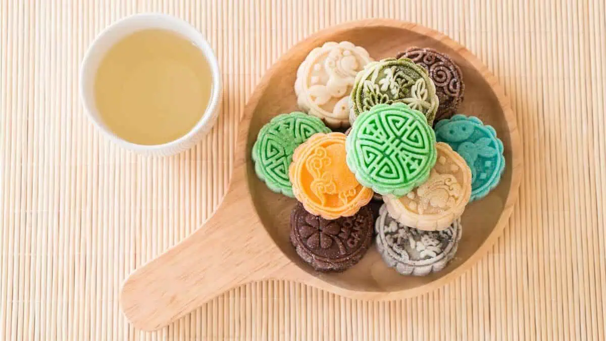 A wooden dish holds colorful, intricately designed mooncakes next to a cup of tea.