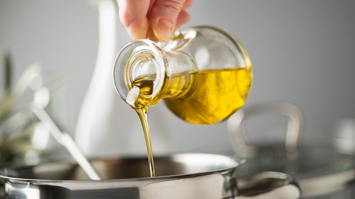A hand pours olive oil from a glass into a cooking pot in a kitchen.