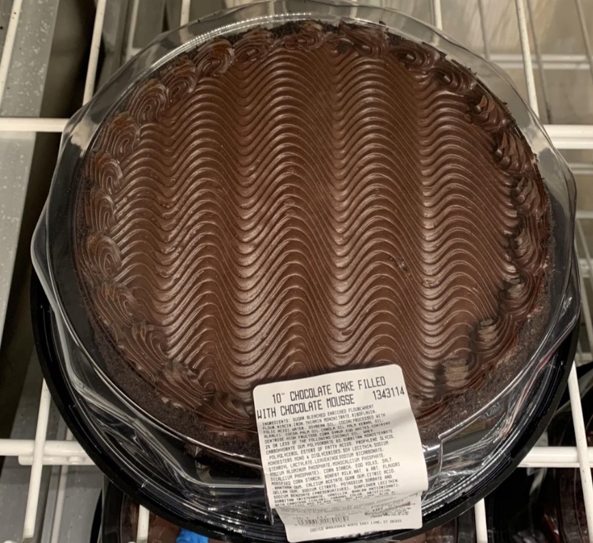 A round chocolate cake with chocolate mousse filling, topped with decorative wavy swirls of chocolate frosting, sits in a clear plastic container with a label on top.