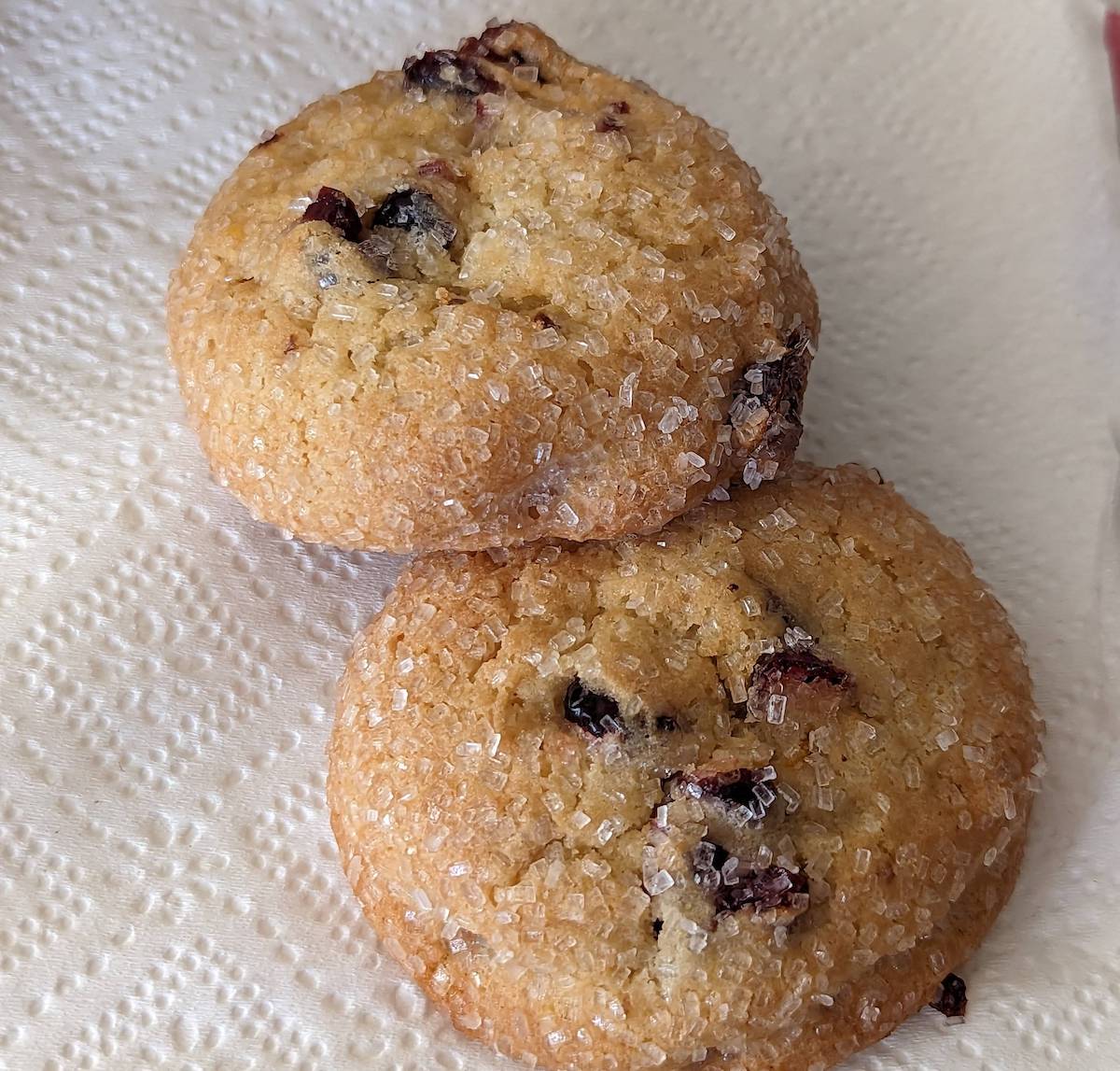 Two golden-brown cookies with visible sugar crystals and bits of dried fruit rest on a textured white paper towel.