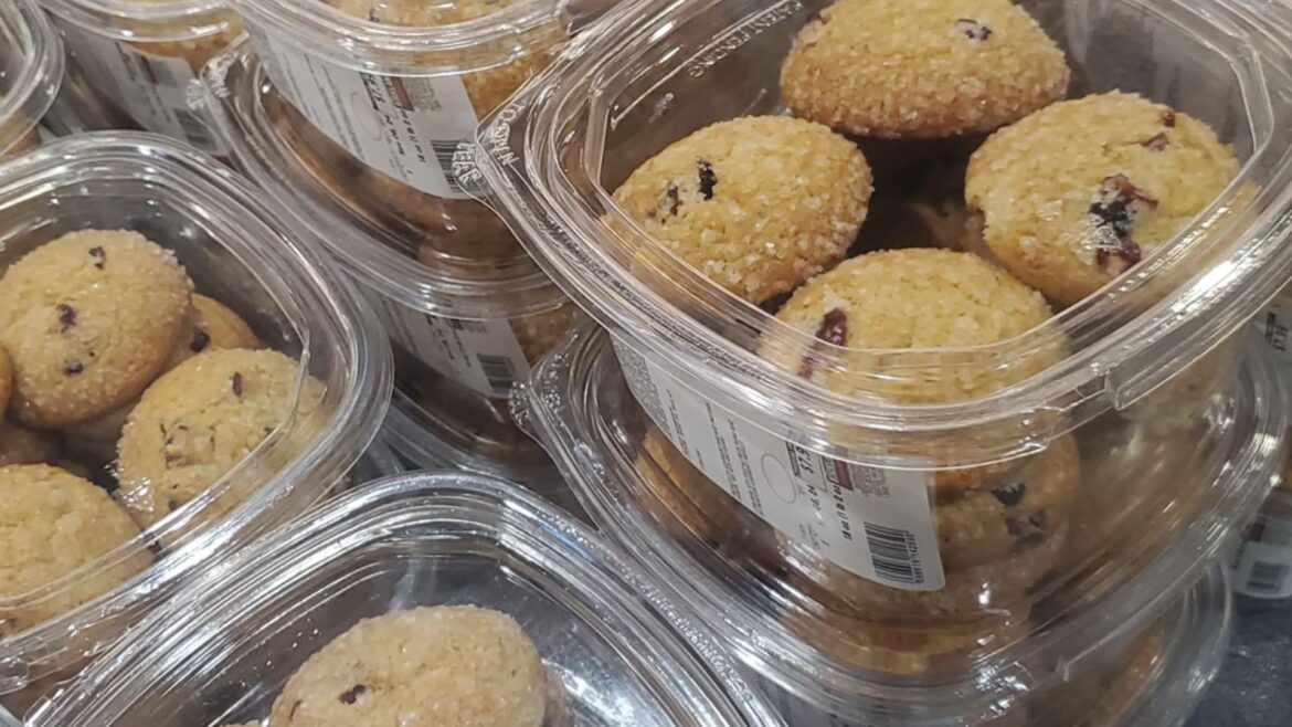 Multiple clear plastic containers filled with sugar-dusted cookies, some with visible chocolate or fruit pieces, are stacked together on display, each labeled with a barcode and text.