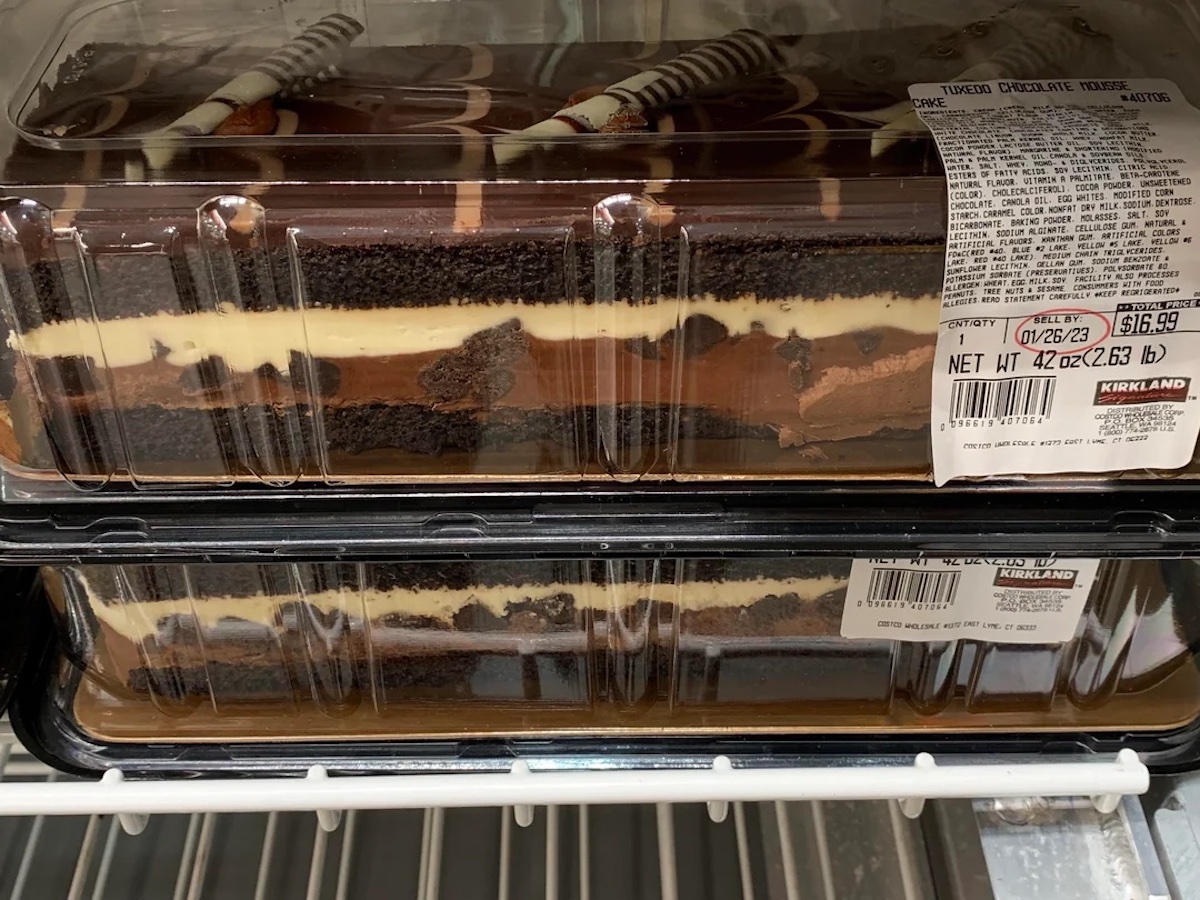 Two clear plastic containers of Kirkland Tuxedo Chocolate Mousse Cake with chocolate, cream, and cake layers are stacked on a refrigerator shelf. The top container has a visible price label.