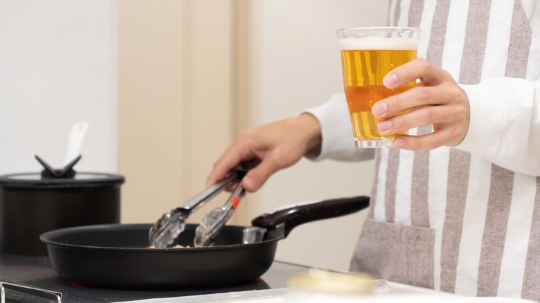 Person holding glass of beer while cooking with frying pan