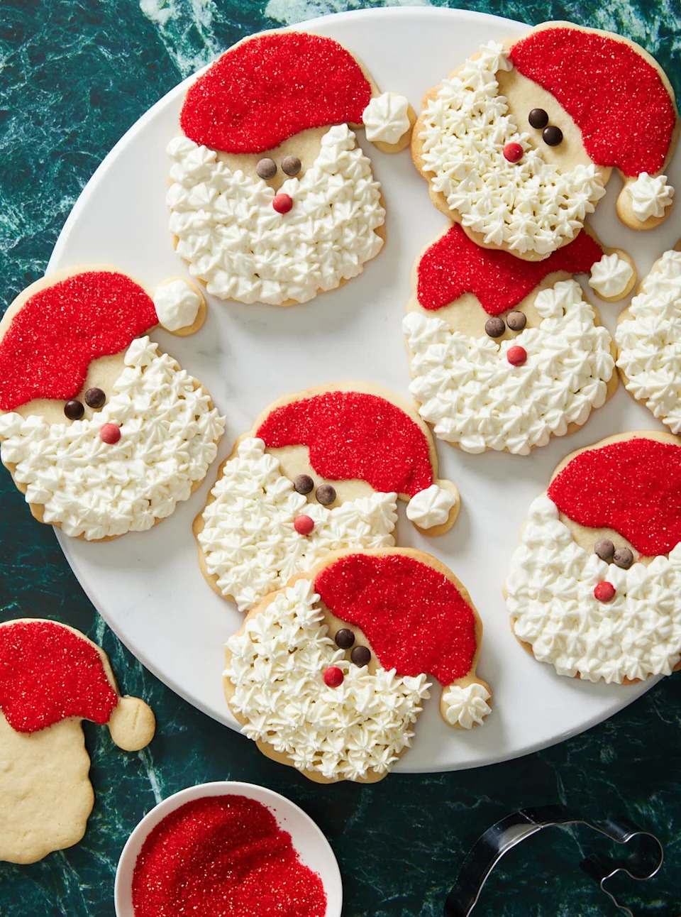 sugar cookies decorated like santa claus with white frosting, red sprinkles, and chocolate for eyes