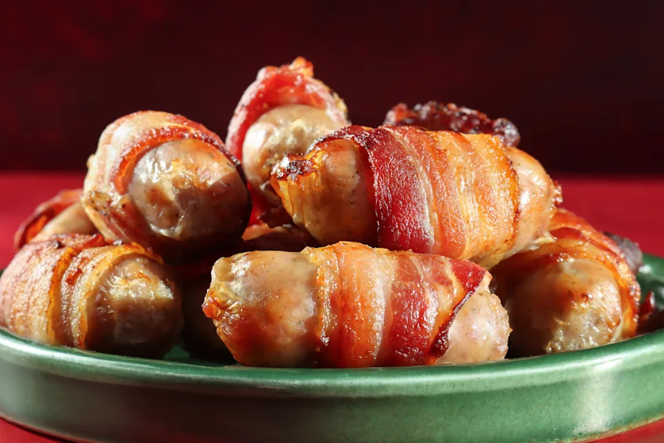 Stock photo shows festive Christmas plate showcasing crispy pigs in blankets, sausages wrapped in streaky bacon, garnished with fresh rosemary and herbs. This savory holiday appetizer features golden-brown, flavorful bites perfect for festive meals and entertaining. The image highlights traditional seasonal comfort food, emphasizing appealing presentation, rich textures, and aromatic garnishes