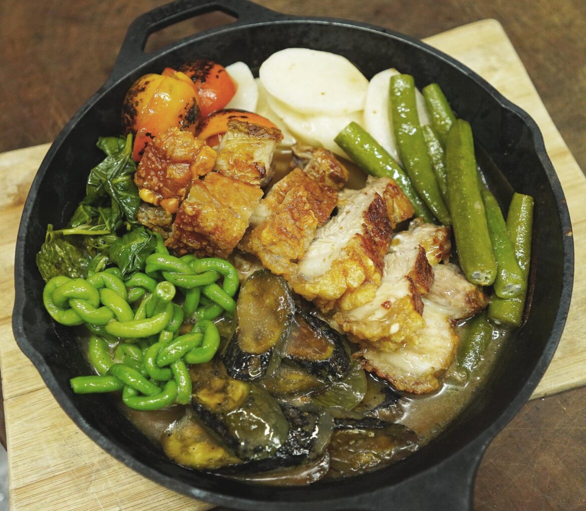 Crispy Pork Kare Kare in a cast-iron skillet golden crackling, rich peanut sauce, charred tomatoes, eggplant, long beans, and greens, styled for peak food photography [OC].