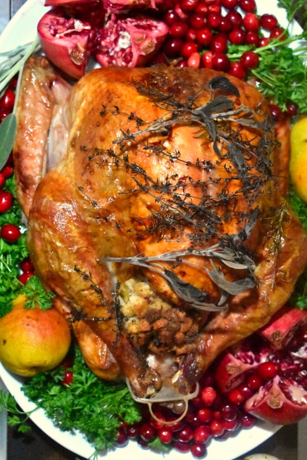 overhead view of roasted turkey on a white platter and garnished with fresh cranberries, sliced pomegranate, pears, sage and parsley
