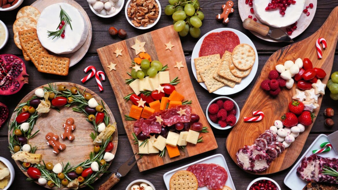 A festive charcuterie spread with meats, cheeses, crackers, fruits, nuts, raspberries, pomegranate, and candies. Boards are decorated with herbs, gingerbread cookies, and holiday-themed garnishes.