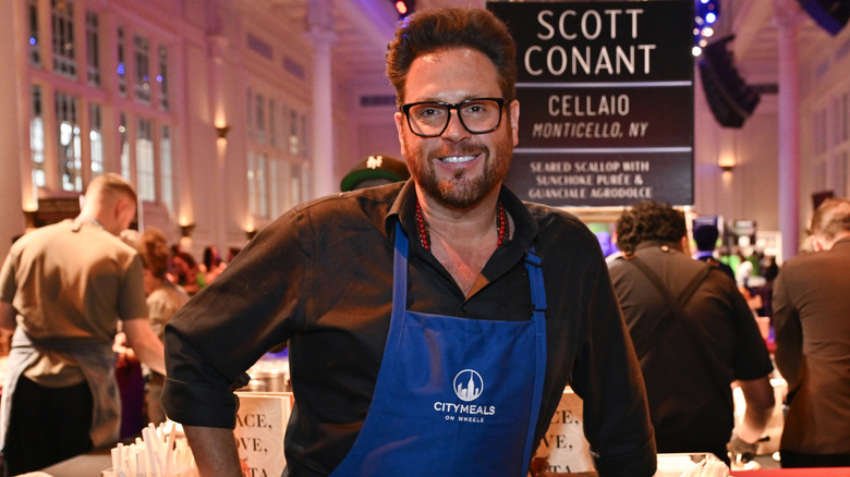 Scott Conant wearing a City Meals On Wheels blue apron and smiling at a food event