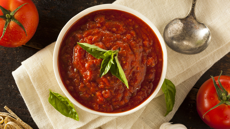 bowl of marinara sauce topped with basil leaves sitting on a linen napkin surrounded by fresh ingredients and spoon