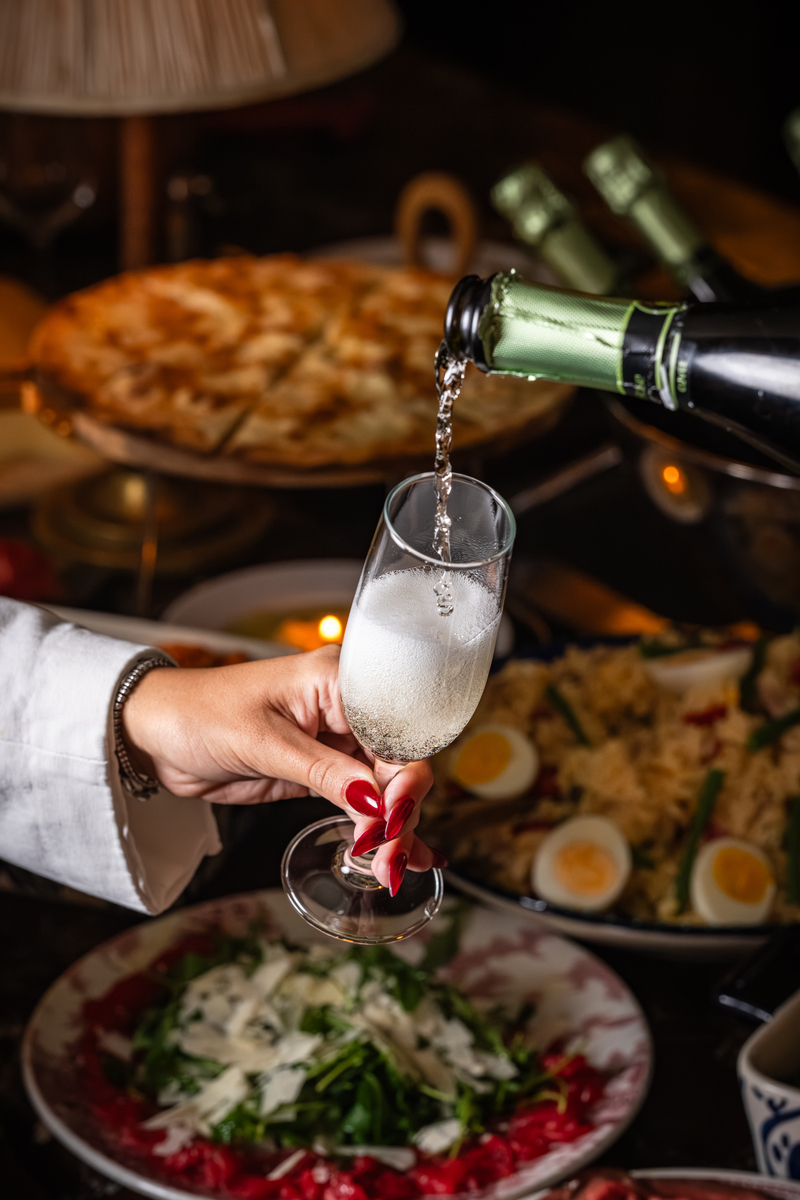prosecco being poured into an outstretched flute, with a feat of italian food visible in the background