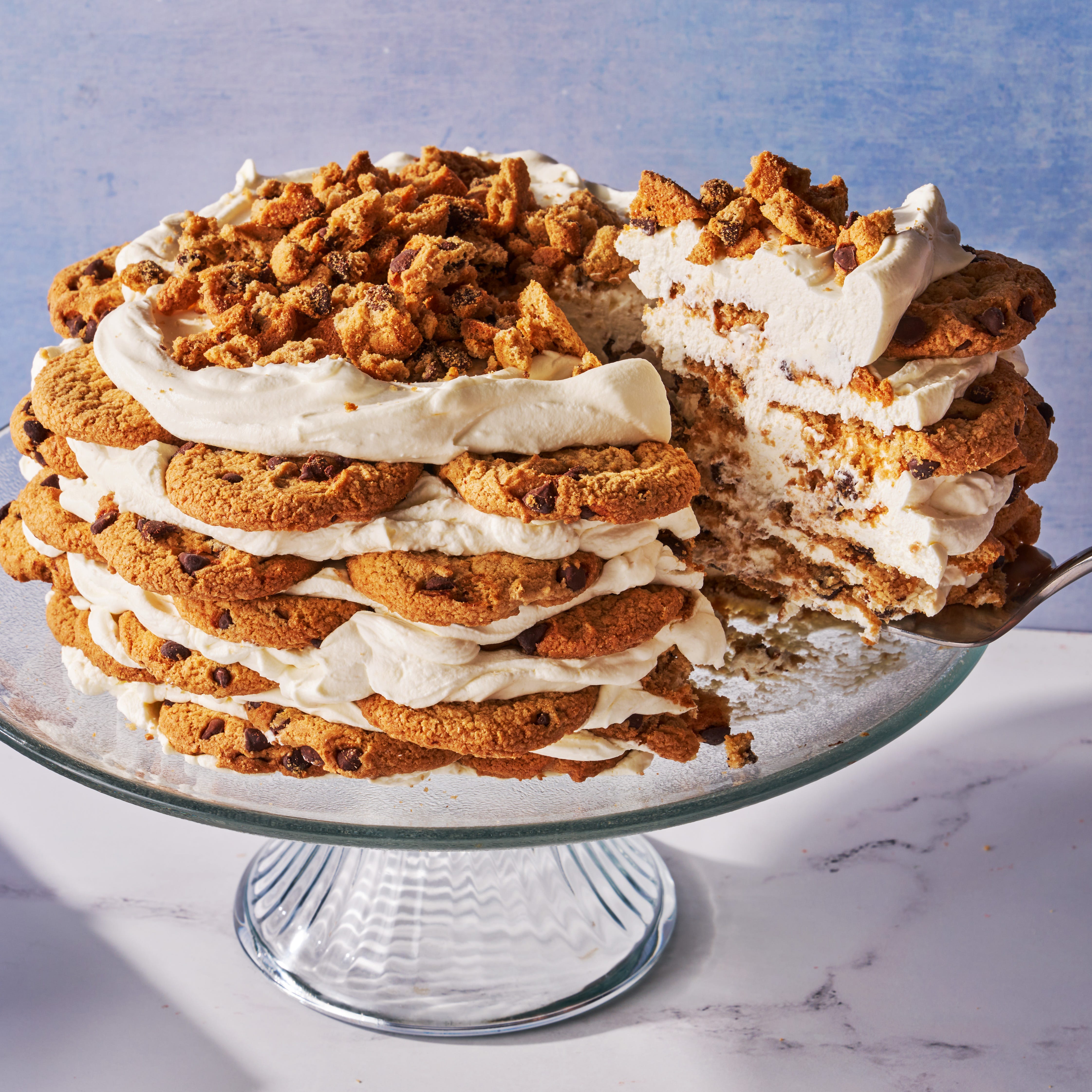 icebox cake made of chocolate chip cookies and whipped cream layered on a cake stand in front of a blue background