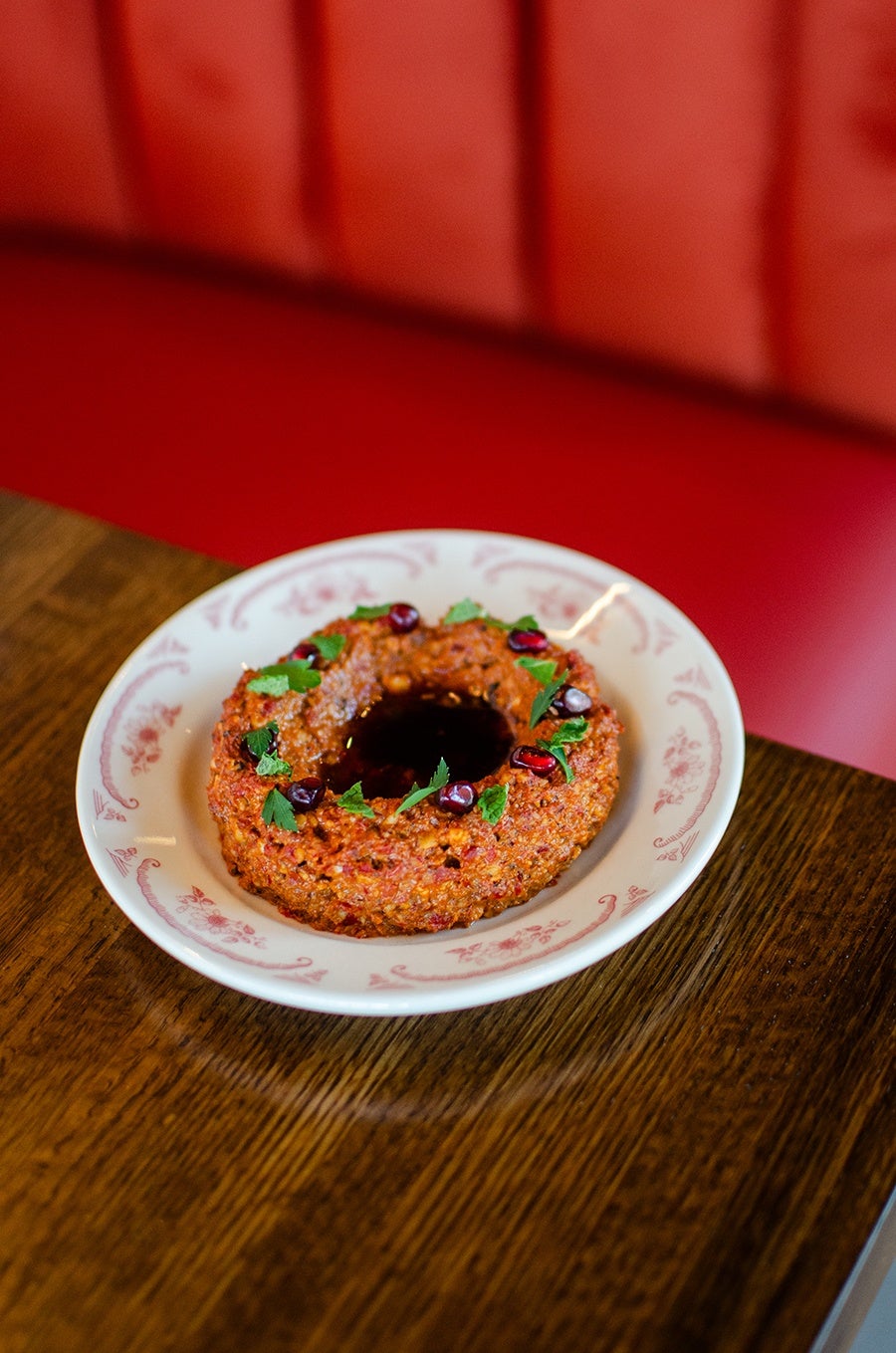 A ring-shaped red dip or spread garnished with fresh green herbs and pomegranate seeds, served on a white plate with a pink floral pattern, placed on a wooden table with a red cushioned background.