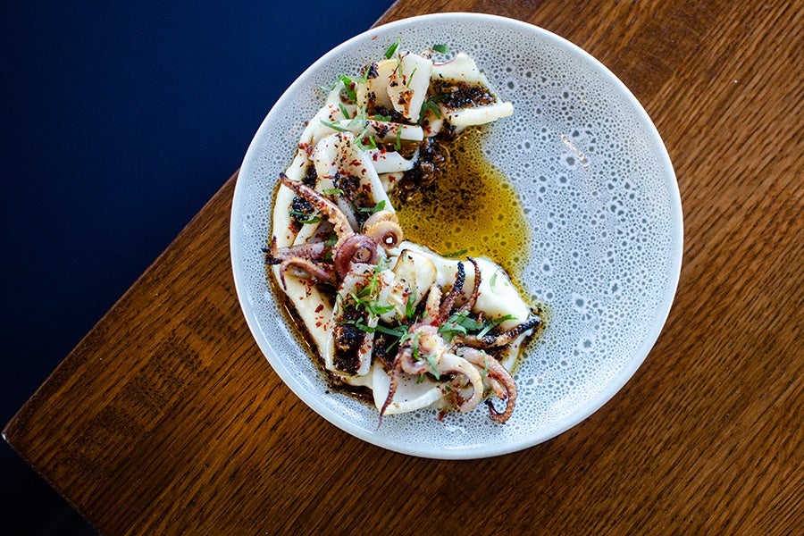 Grilled squid tentacles served on a white plate with a textured pattern, garnished with chopped herbs and spices, and drizzled with olive oil, placed on a wooden table.