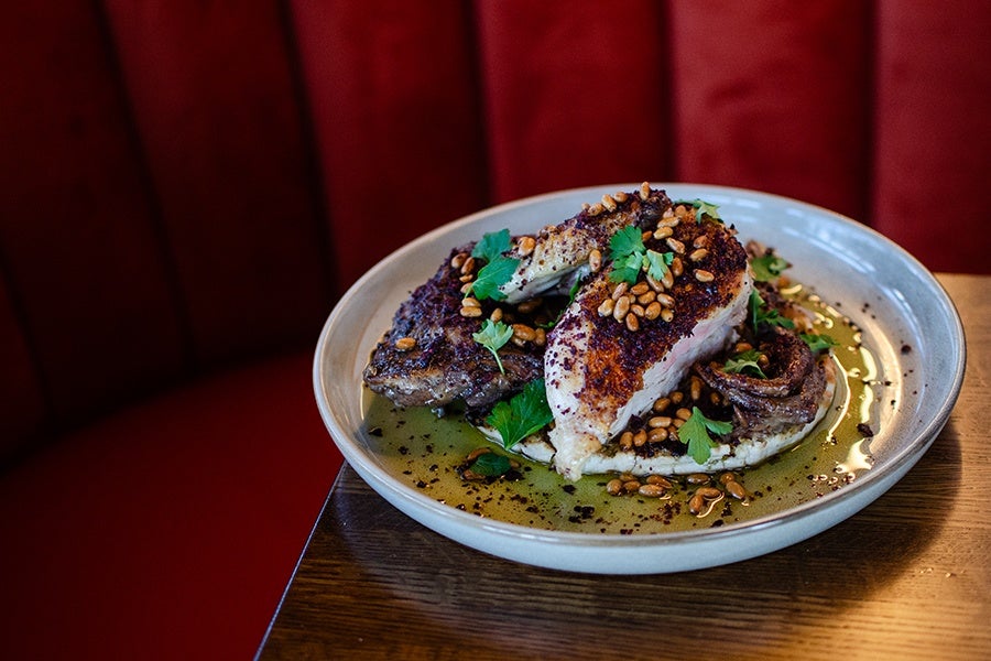 A plate of roasted chicken seasoned with spices, garnished with pine nuts and fresh parsley, served on a bed of creamy mash with a drizzle of olive oil. The dish is presented on a light-colored ceramic plate placed on a wooden table with a red cushioned background.