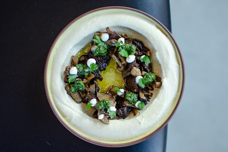 A bowl of creamy hummus topped with sautéed mixed mushrooms, dollops of green herb pesto, small white cream drops, and garnished with fresh parsley leaves. The hummus has a smooth texture with a pool of olive oil in the center.